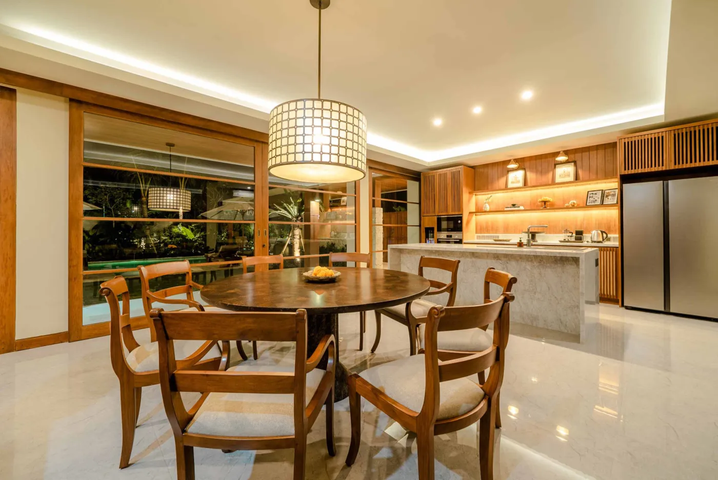 Modern tropical dining area with wooden round table and chairs under pendant light, adjacent open kitchen, and large windows to garden at dusk. - Bali Villas