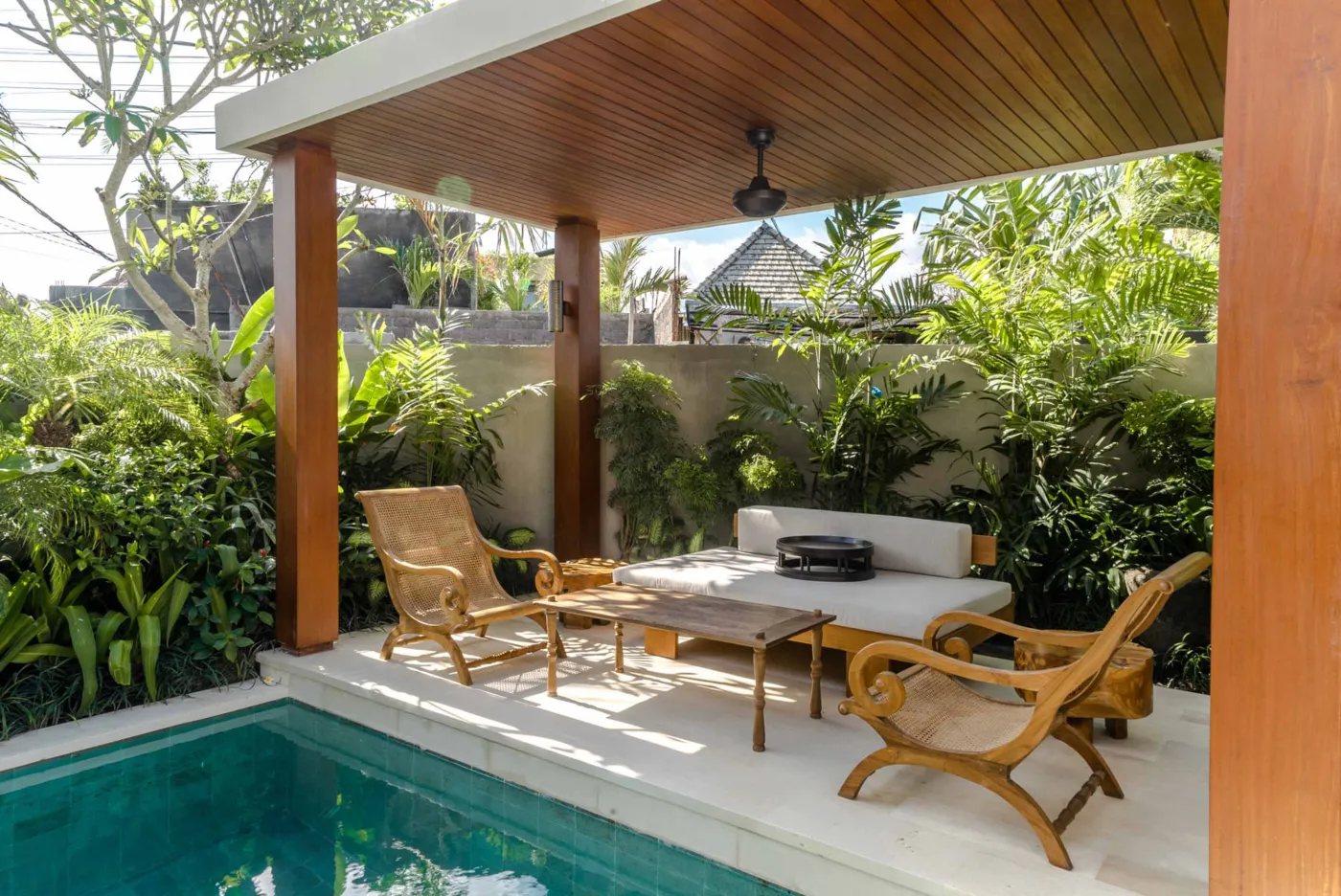 Modern tropical dining area with wooden round table and chairs under pendant light, adjacent open kitchen, and large windows to garden at dusk. - Bali Villas