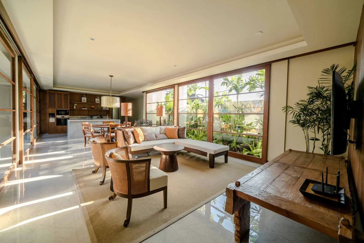 Modern tropical dining area with wooden round table and chairs under pendant light, adjacent open kitchen, and large windows to garden at dusk. - Bali Villas