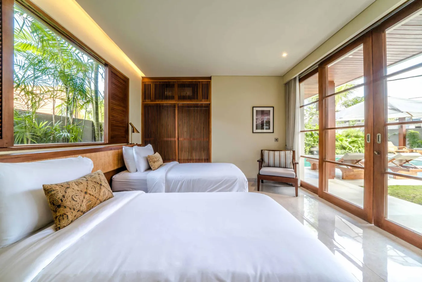 Modern tropical dining area with wooden round table and chairs under pendant light, adjacent open kitchen, and large windows to garden at dusk. - Bali Villas