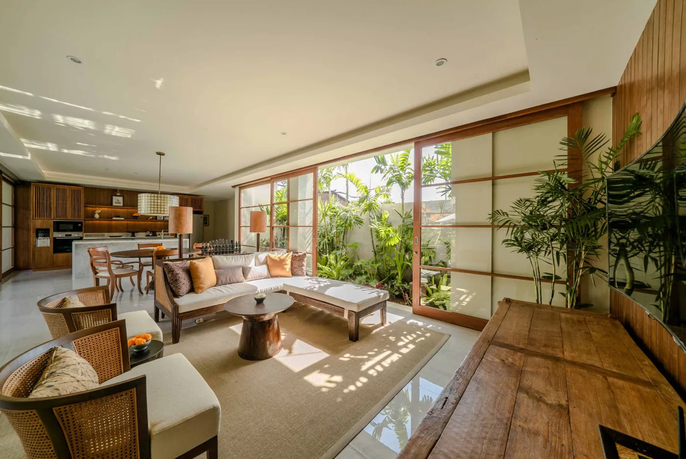 Modern tropical dining area with wooden round table and chairs under pendant light, adjacent open kitchen, and large windows to garden at dusk. - Bali Villas