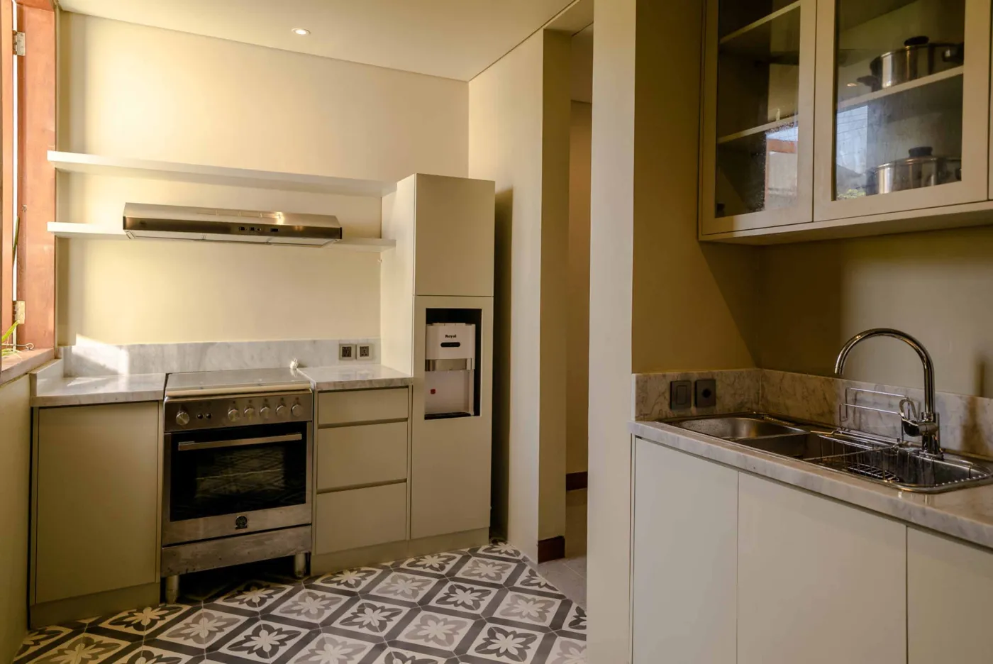 Modern tropical dining area with wooden round table and chairs under pendant light, adjacent open kitchen, and large windows to garden at dusk. - Bali Villas