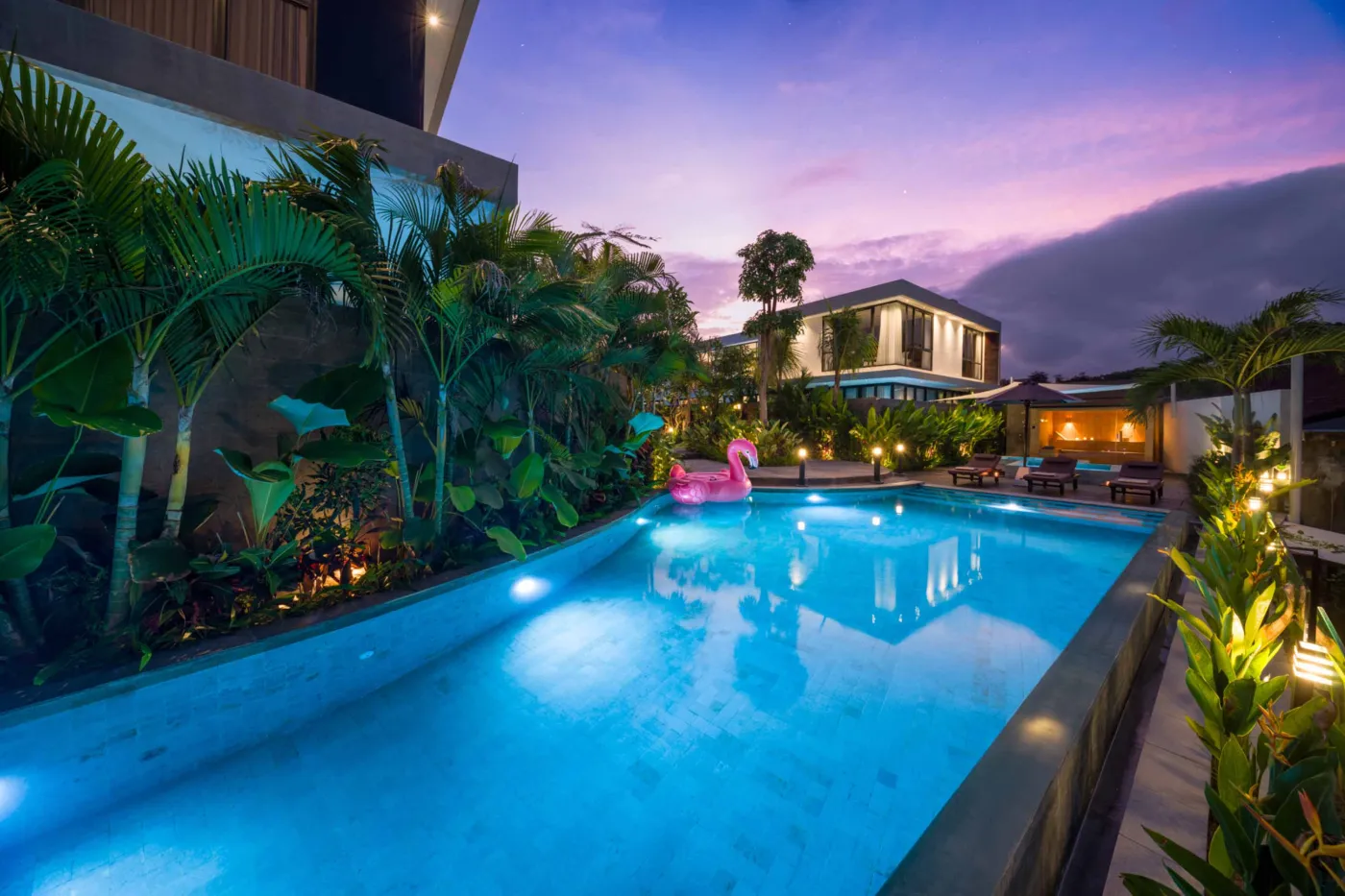 Luxurious tropical resort pool at twilight, featuring a glowing blue infinity pool with pink flamingo float, modern villas, palm trees, and ambient lighting. - Bali Villas