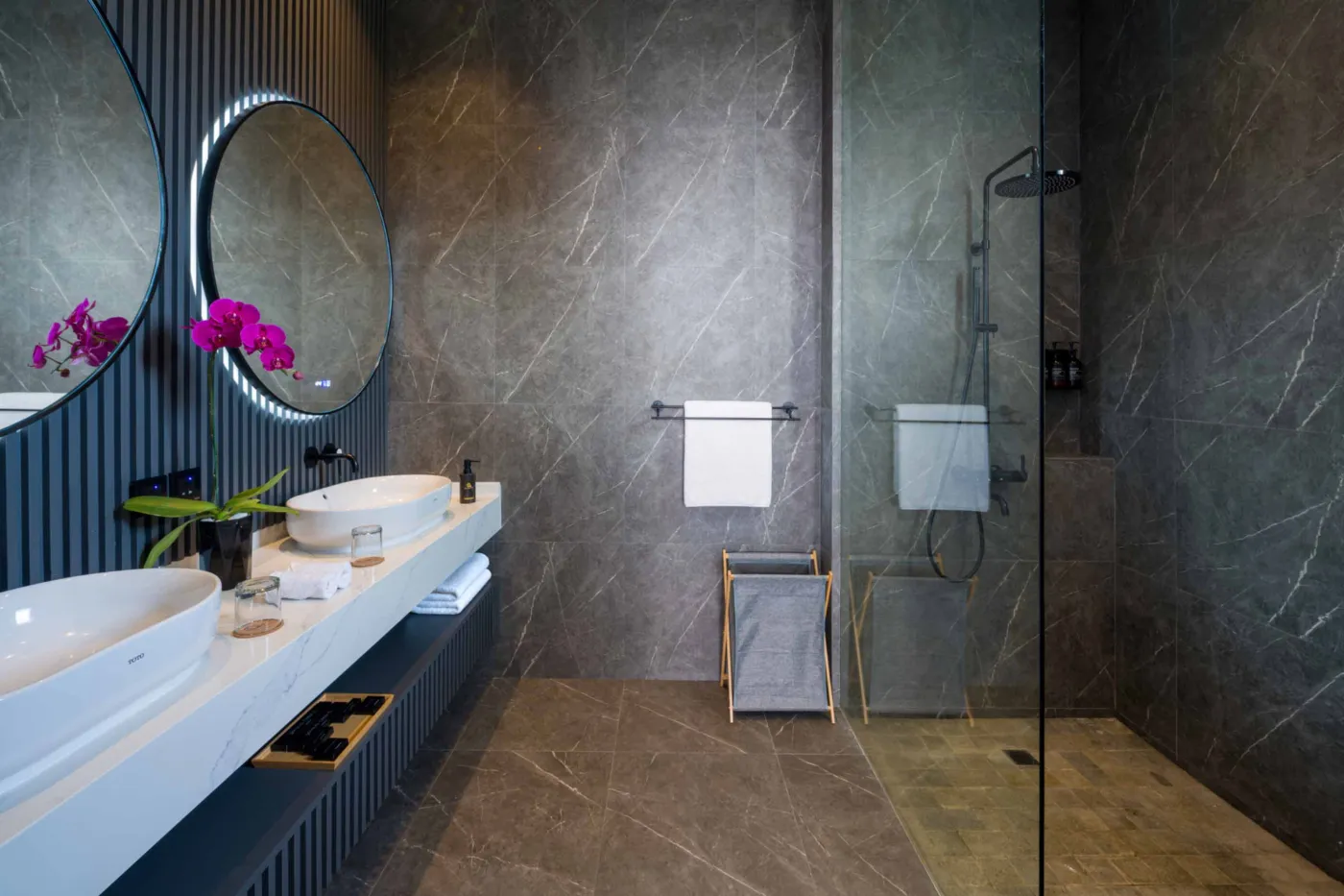 Modern luxury bathroom with gray stone walls, walk-in glass shower, freestanding white vanity, wall-hung toilet, oval backlit mirror, and purple floral accents. - Bali Villas