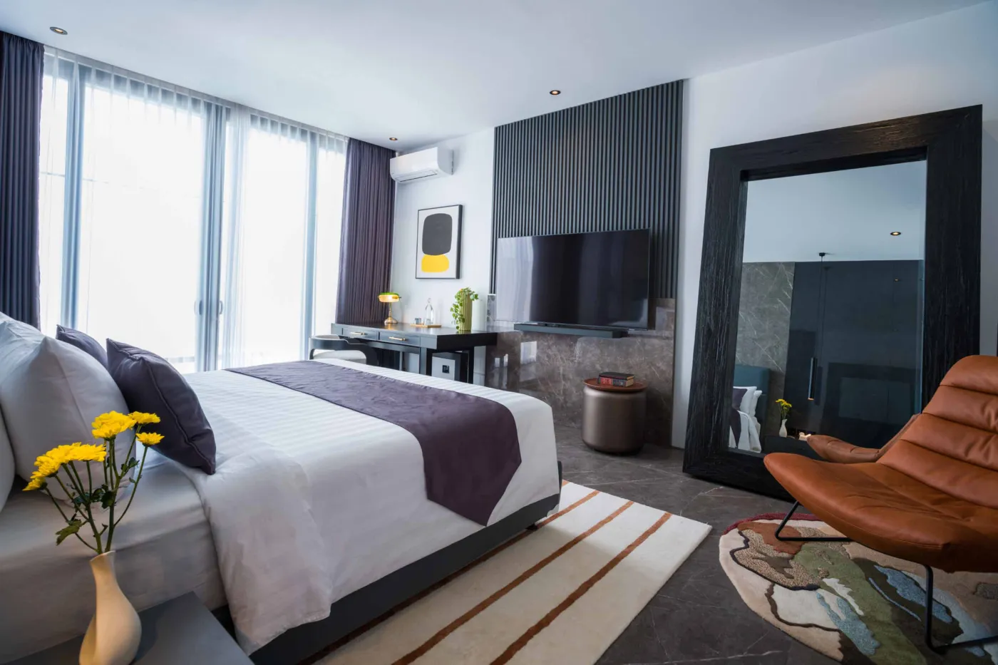 Modern luxury bathroom with gray stone walls, walk-in glass shower, freestanding white vanity, wall-hung toilet, oval backlit mirror, and purple floral accents. - Bali Villas