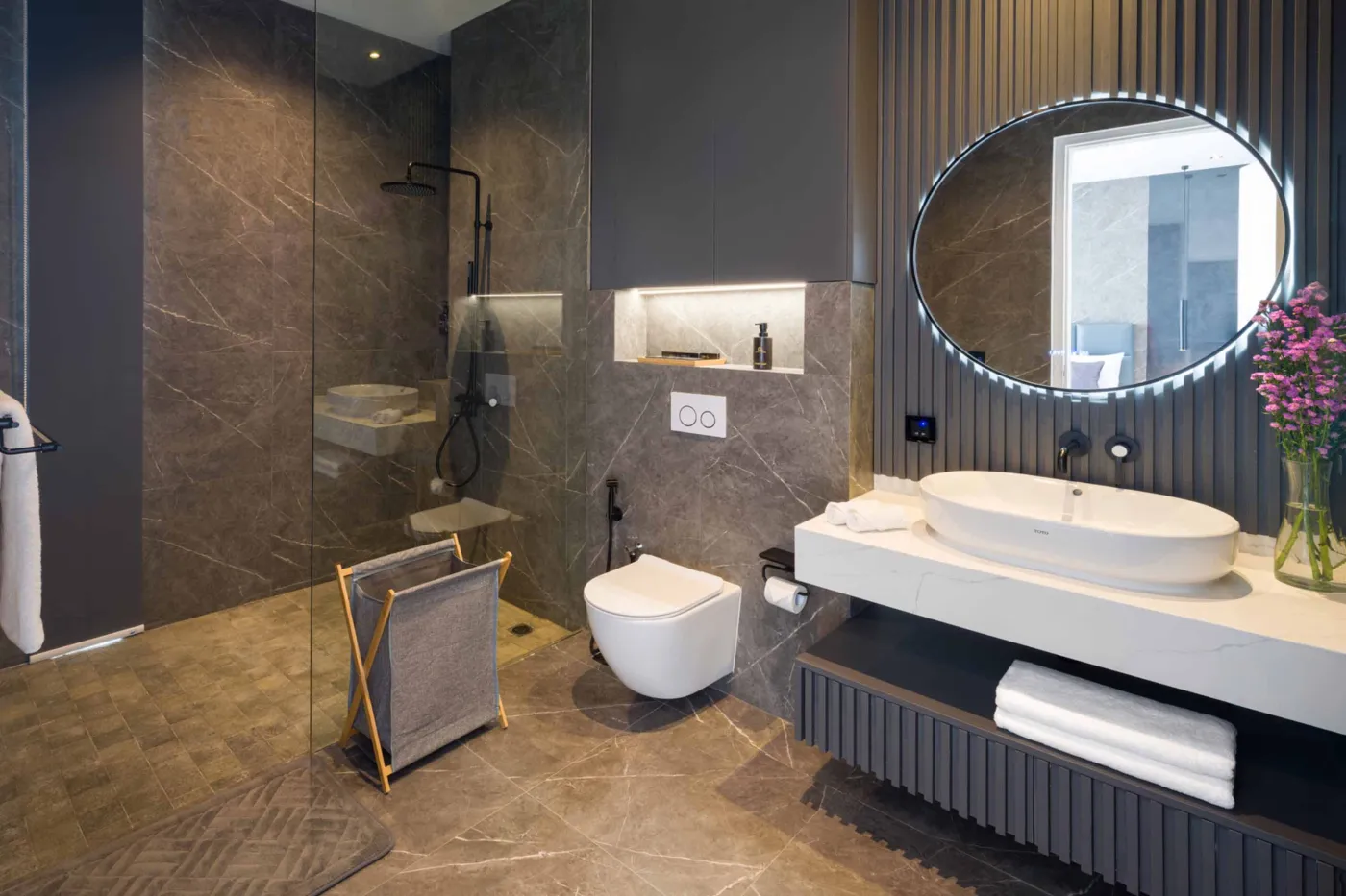 Modern luxury bathroom with gray stone walls, walk-in glass shower, freestanding white vanity, wall-hung toilet, oval backlit mirror, and purple floral accents. - Bali Villas