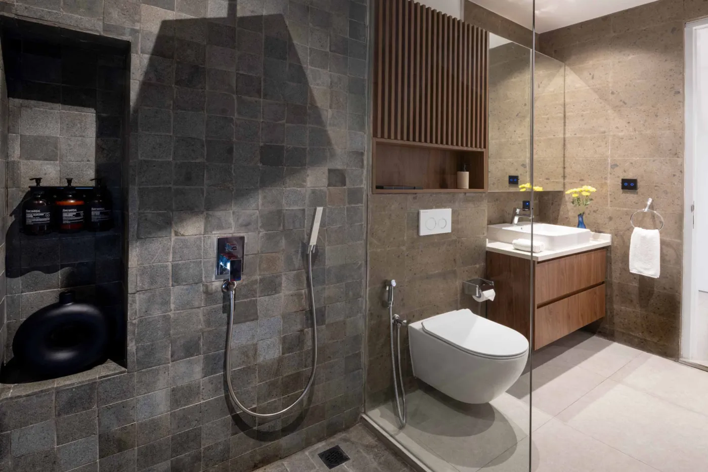 Modern minimalist bathroom with gray tiled walk-in shower, wooden slatted accents, wall-mounted toilet, and sleek vanity sink. - Bali Villas