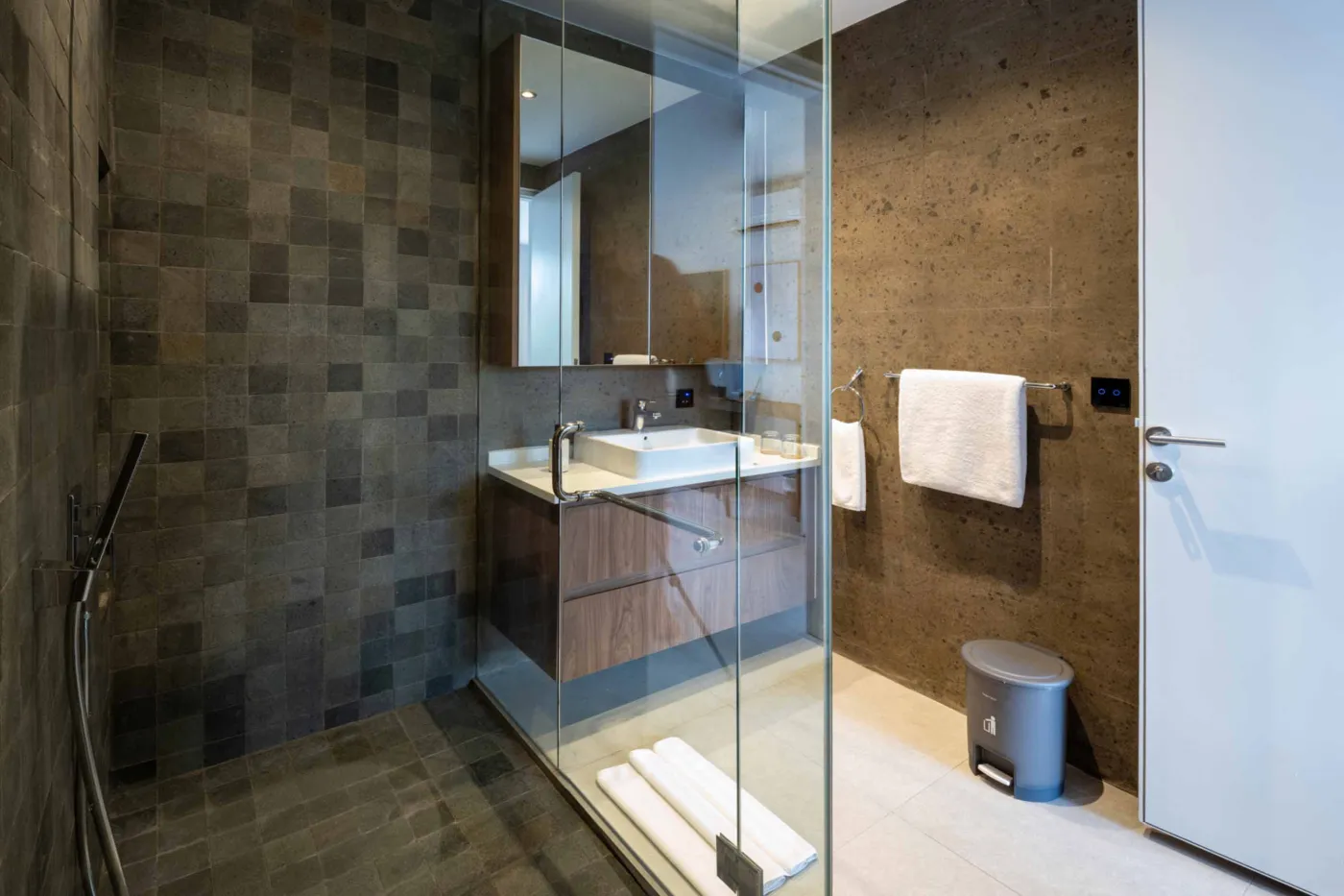 Modern minimalist bathroom with gray tiled walk-in shower, wooden slatted accents, wall-mounted toilet, and sleek vanity sink. - Bali Villas