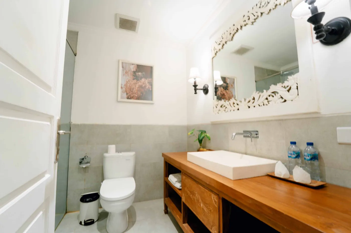 Modern bathroom with wooden vanity, white sink, ornate mirror, wall sconces, potted plant, toilet, and bottled water. - Bali Villas