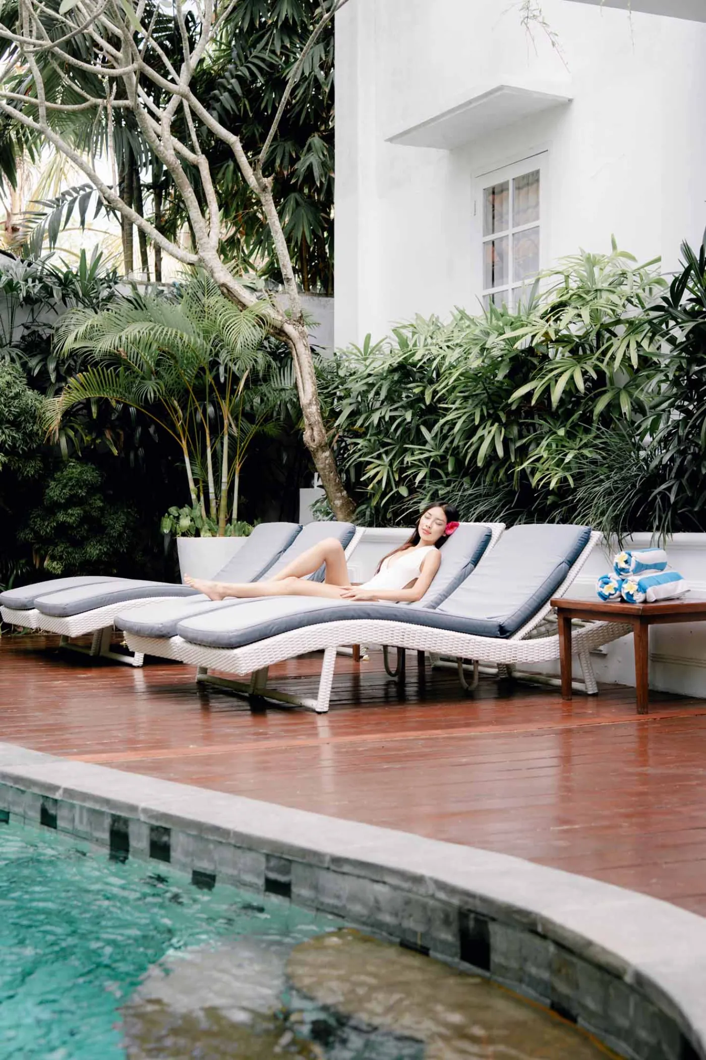 Woman lounging on a poolside chaise in a tropical resort, surrounded by palm trees, white villa, and turquoise infinity pool. - Bali Villas
