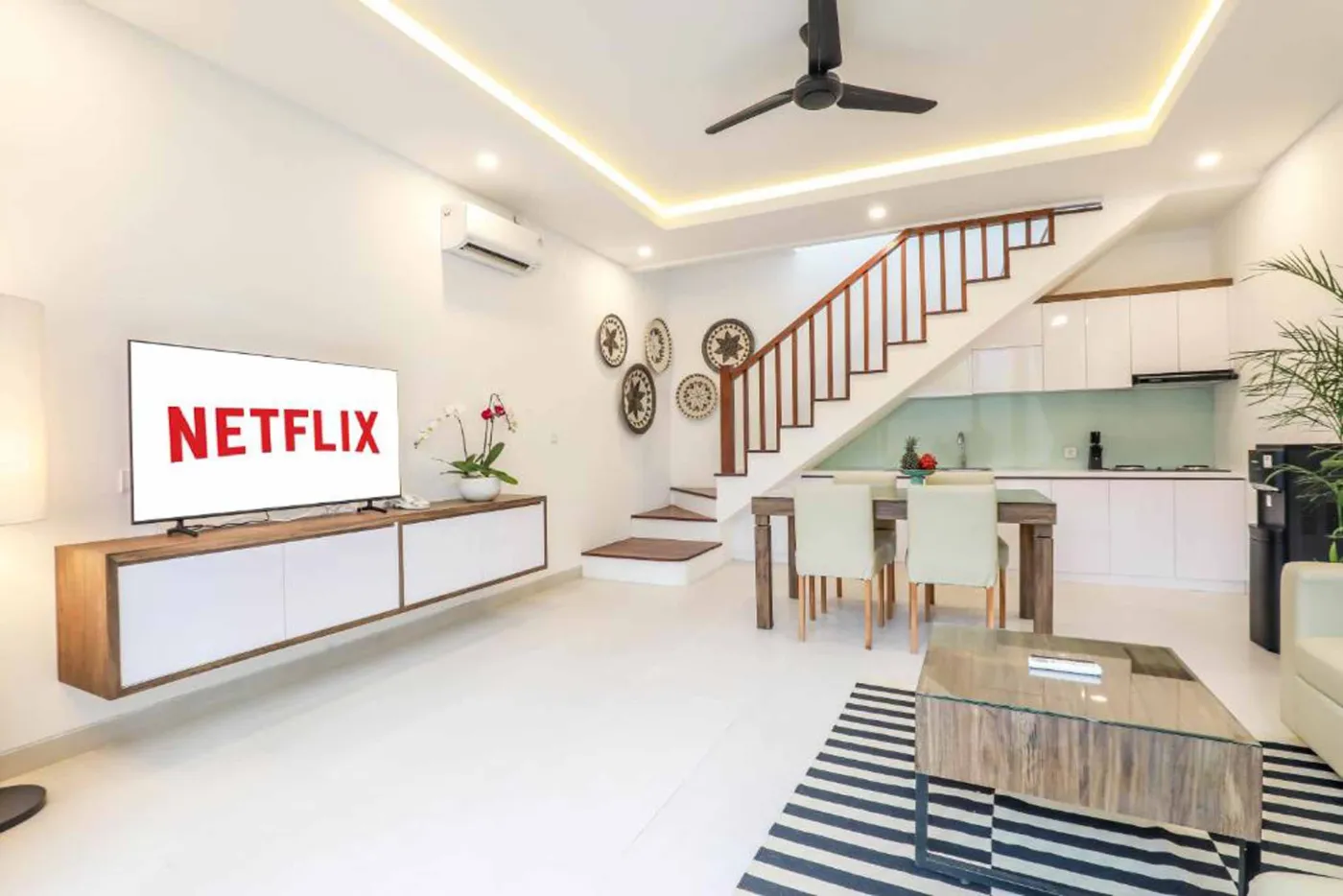 Modern Bali villa living room with beige sofas, glass dining table, Netflix on wall TV, potted plants, and sliding doors to infinity pool. - Bali Villas