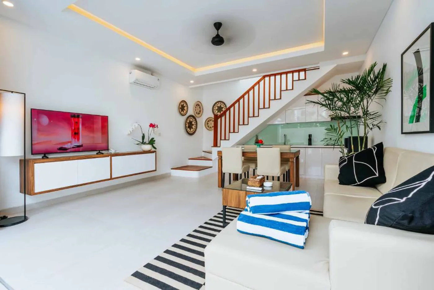 Modern Bali villa living room with beige sofas, glass dining table, Netflix on wall TV, potted plants, and sliding doors to infinity pool. - Bali Villas