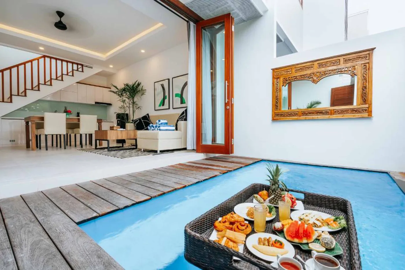Modern Bali villa living room with beige sofas, glass dining table, Netflix on wall TV, potted plants, and sliding doors to infinity pool. - Bali Villas