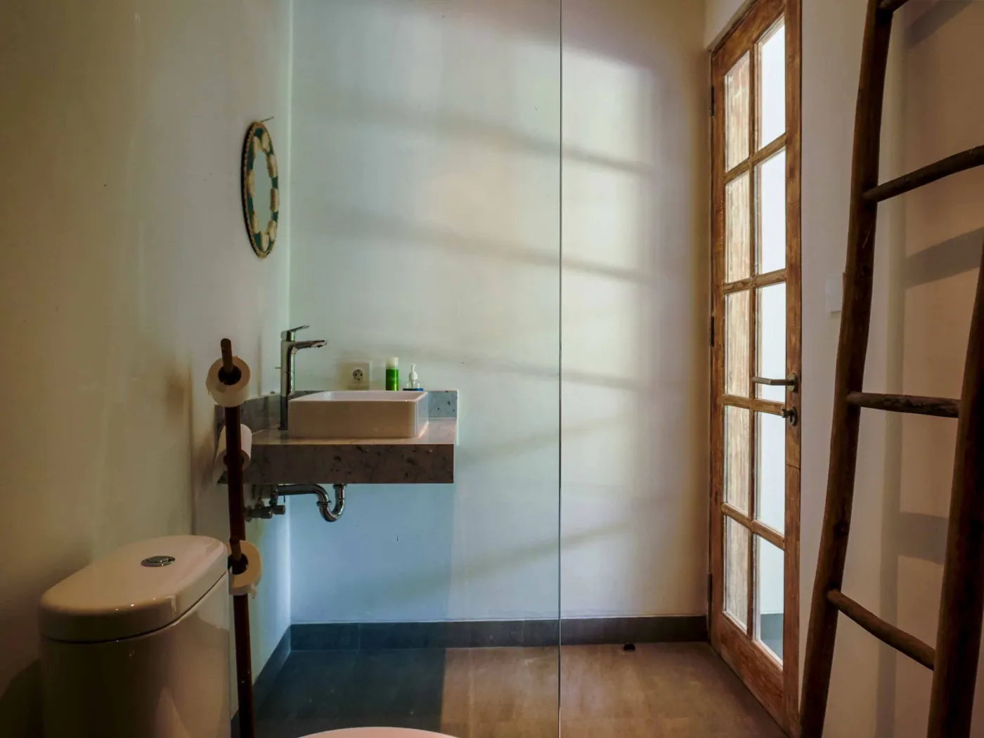 Minimalist modern bathroom with open wooden French door, wall-mounted white sink, round mirror, wooden towel rack, and corner toilet. - Bali Villas