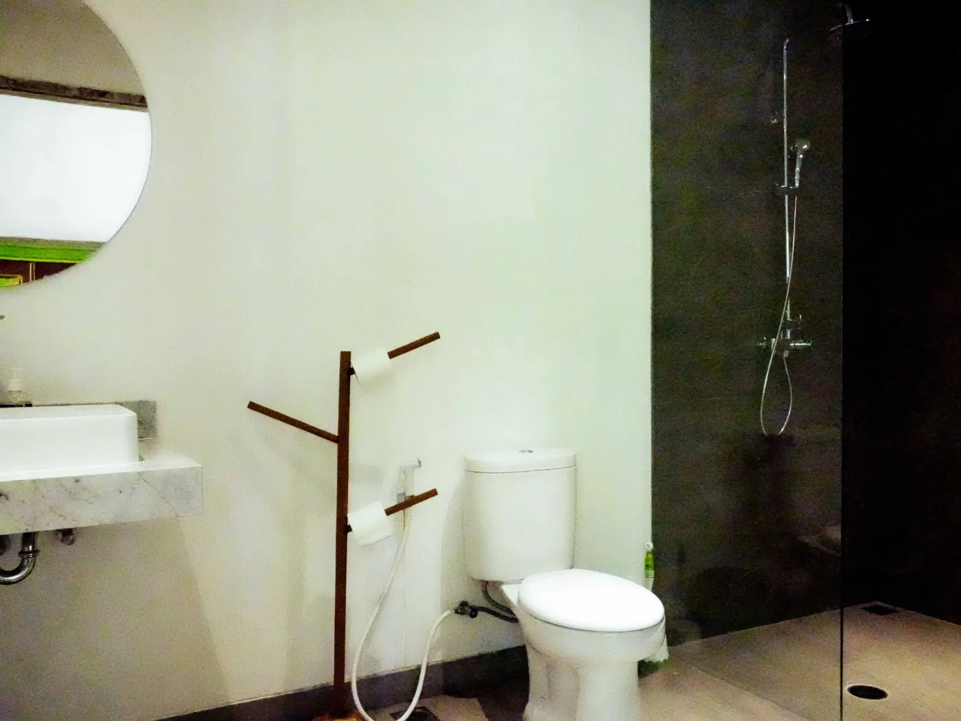 Minimalist modern bathroom with open wooden French door, wall-mounted white sink, round mirror, wooden towel rack, and corner toilet. - Bali Villas