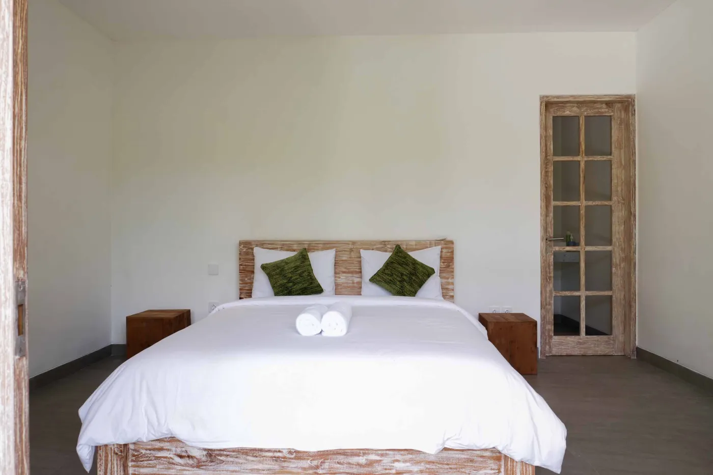 Minimalist modern bathroom with open wooden French door, wall-mounted white sink, round mirror, wooden towel rack, and corner toilet. - Bali Villas