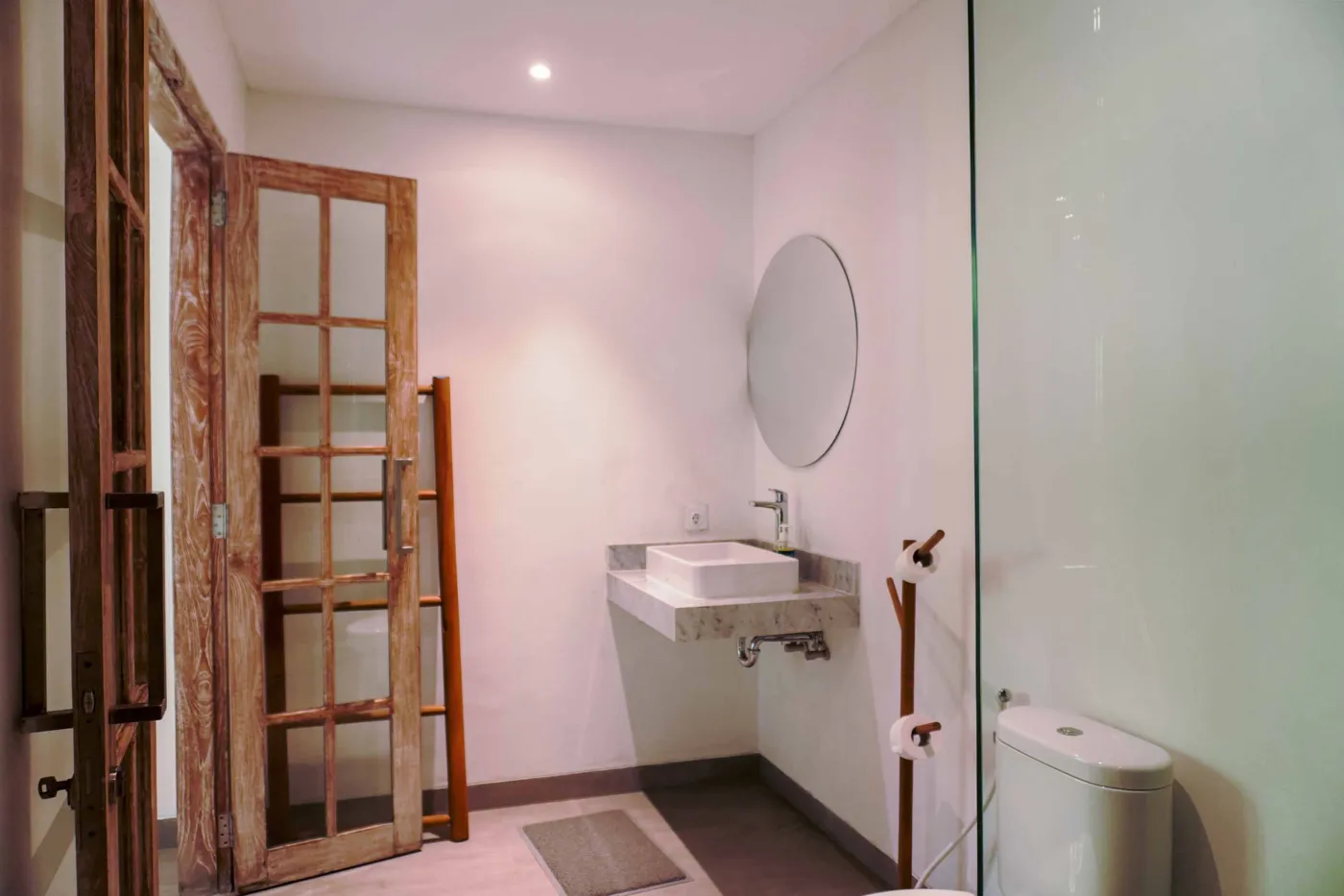 Minimalist modern bathroom with open wooden French door, wall-mounted white sink, round mirror, wooden towel rack, and corner toilet. - Bali Villas