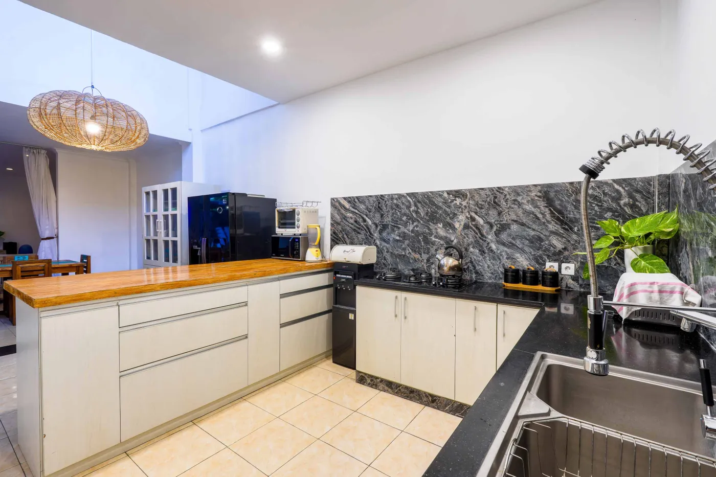Modern white kitchen featuring wooden island, black granite counters, pull-out faucet sink, and woven pendant light. - Bali Villas