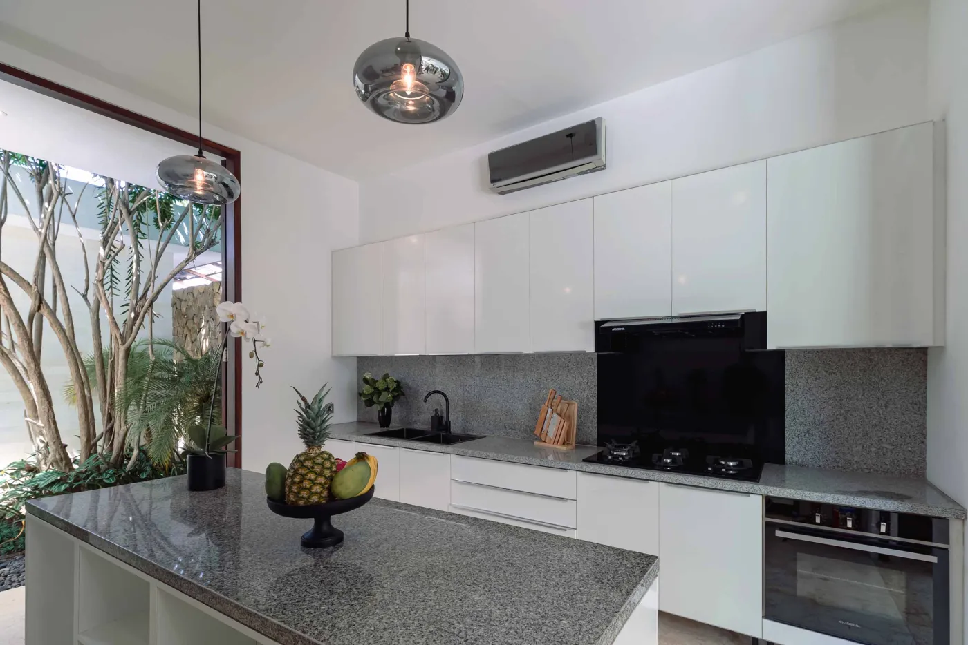 Modern tropical kitchen with white cabinets, granite island topped with fresh fruits, black appliances, and views of lush greenery. - Bali Villas
