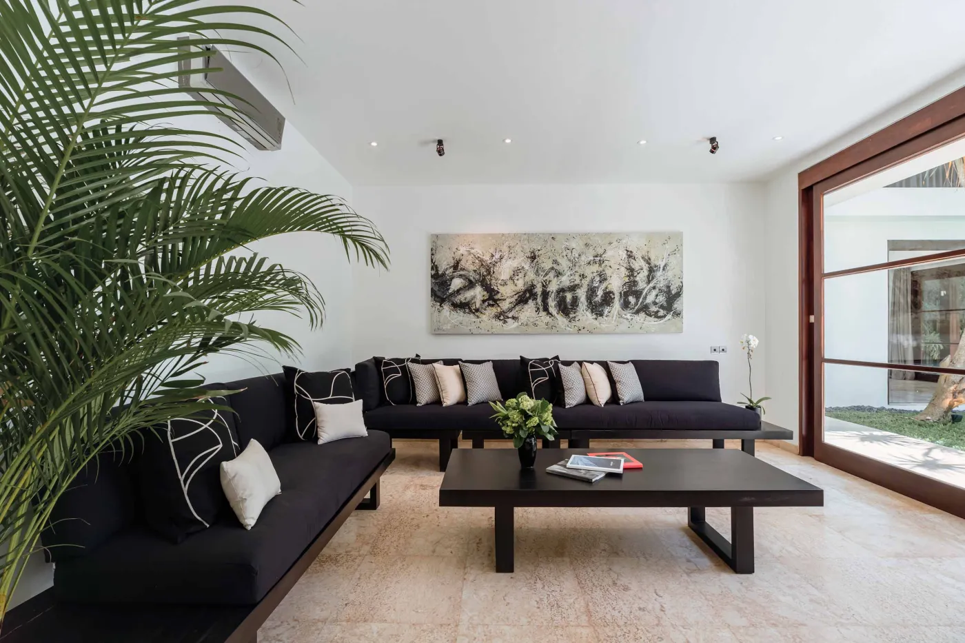 Modern tropical kitchen with white cabinets, granite island topped with fresh fruits, black appliances, and views of lush greenery. - Bali Villas