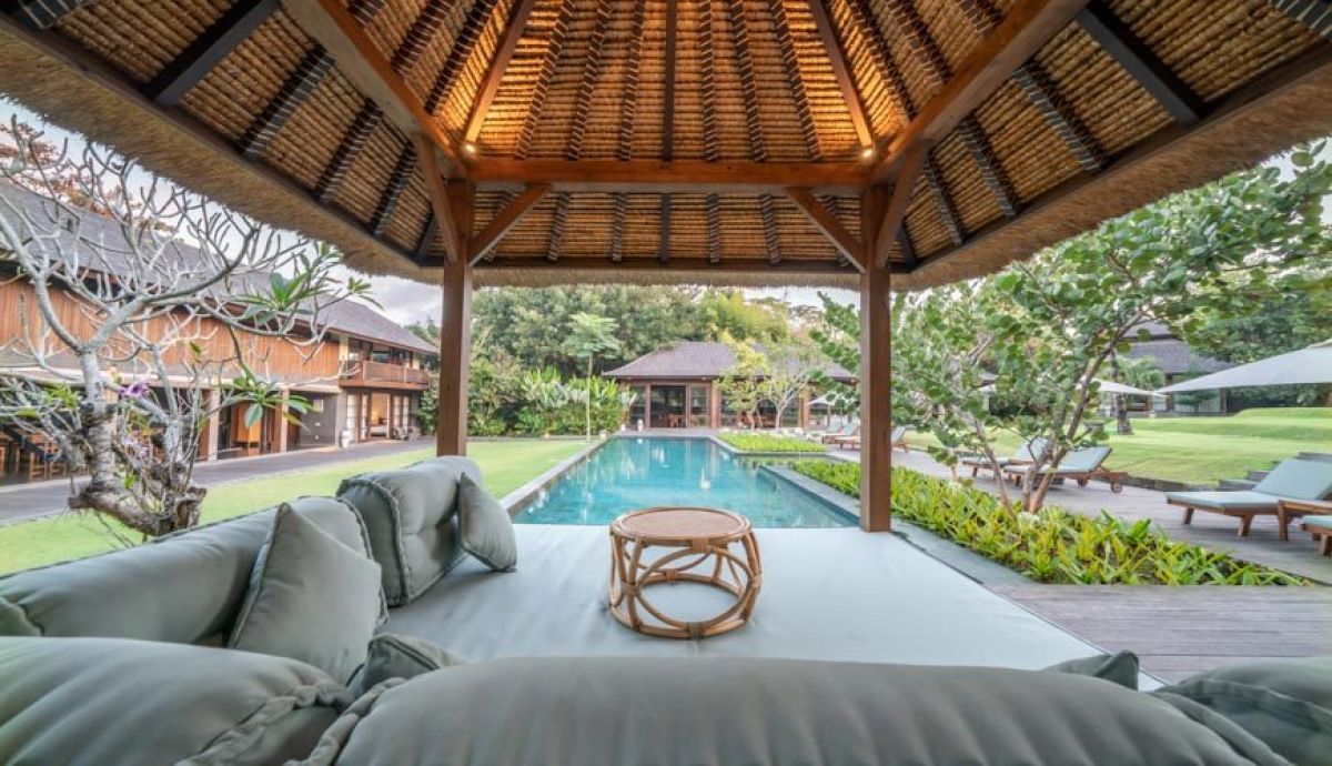 Luxurious open-air Balinese pavilion with lounge seating beside a turquoise infinity pool in a tropical resort garden. - Bali Villas