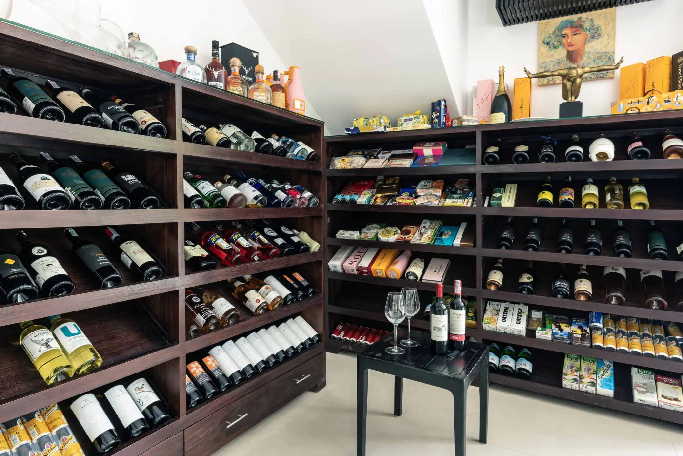 Well-stocked wooden wine cellar featuring diverse bottles of wine and spirits on shelves, with a small tasting table holding glasses and a red wine bottle. - Bali Villas