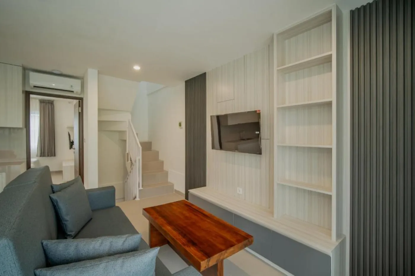 Modern minimalist living room interior with gray sofa, wooden coffee table, wall-mounted TV, built-in white shelves, and adjacent stairs. - Bali Villas