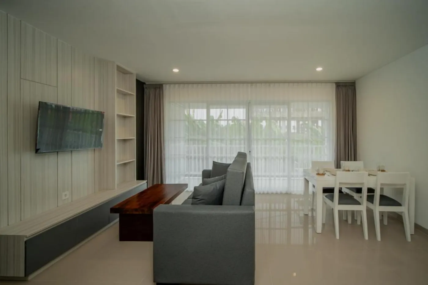 Modern minimalist living room interior with gray sofa, wooden coffee table, wall-mounted TV, built-in white shelves, and adjacent stairs. - Bali Villas