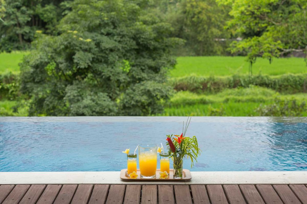 Spacious Balinese villa living room with wooden balcony, tropical furniture, stone statues, and sliding doors to lush garden. - Bali Villas