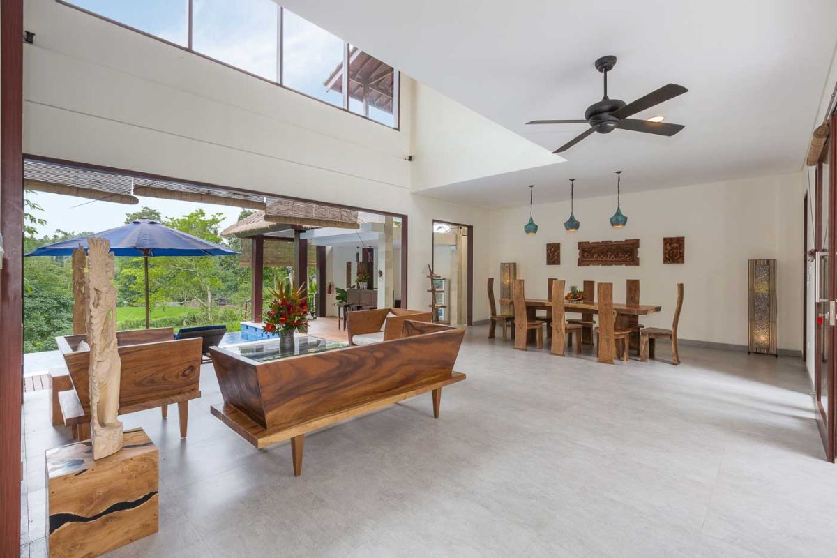 Spacious Balinese villa living room with wooden balcony, tropical furniture, stone statues, and sliding doors to lush garden. - Bali Villas