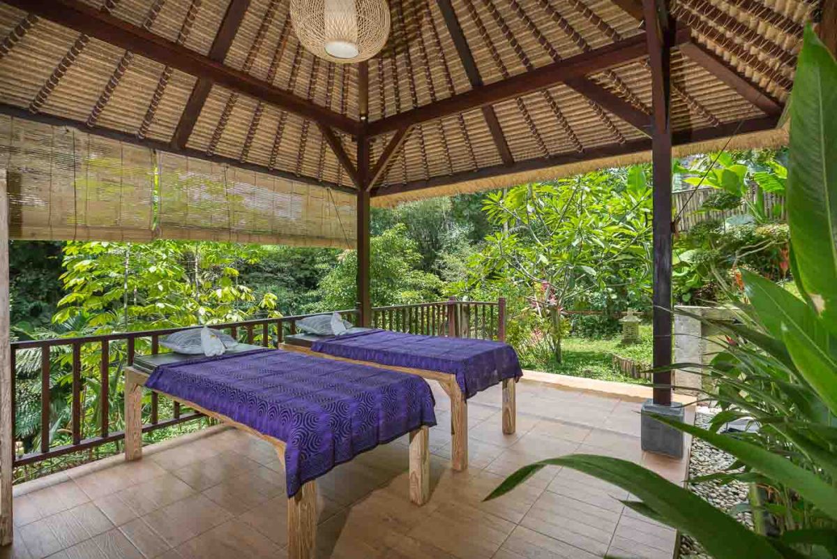 Spacious Balinese villa living room with wooden balcony, tropical furniture, stone statues, and sliding doors to lush garden. - Bali Villas