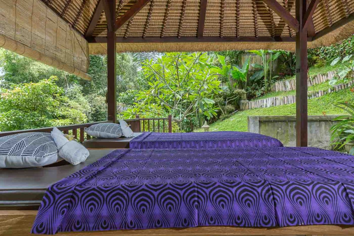 Spacious Balinese villa living room with wooden balcony, tropical furniture, stone statues, and sliding doors to lush garden. - Bali Villas