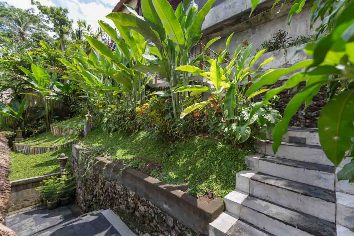 Spacious Balinese villa living room with wooden balcony, tropical furniture, stone statues, and sliding doors to lush garden. - Bali Villas
