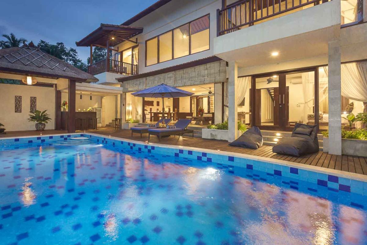 Spacious Balinese villa living room with wooden balcony, tropical furniture, stone statues, and sliding doors to lush garden. - Bali Villas