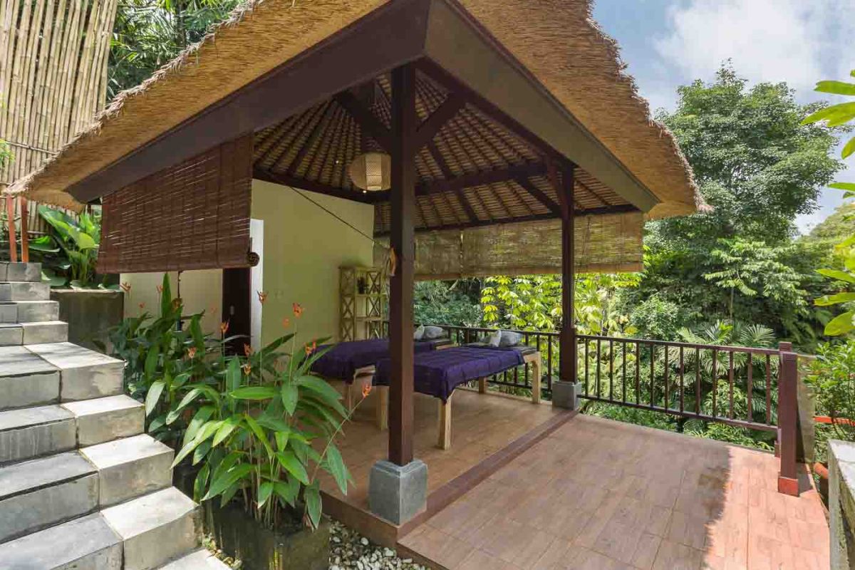 Spacious Balinese villa living room with wooden balcony, tropical furniture, stone statues, and sliding doors to lush garden. - Bali Villas