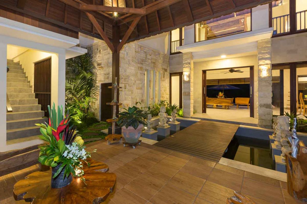Spacious Balinese villa living room with wooden balcony, tropical furniture, stone statues, and sliding doors to lush garden. - Bali Villas
