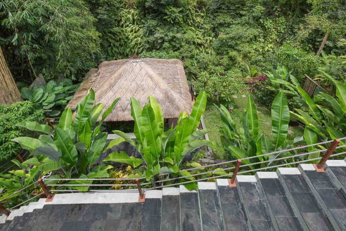 Spacious Balinese villa living room with wooden balcony, tropical furniture, stone statues, and sliding doors to lush garden. - Bali Villas