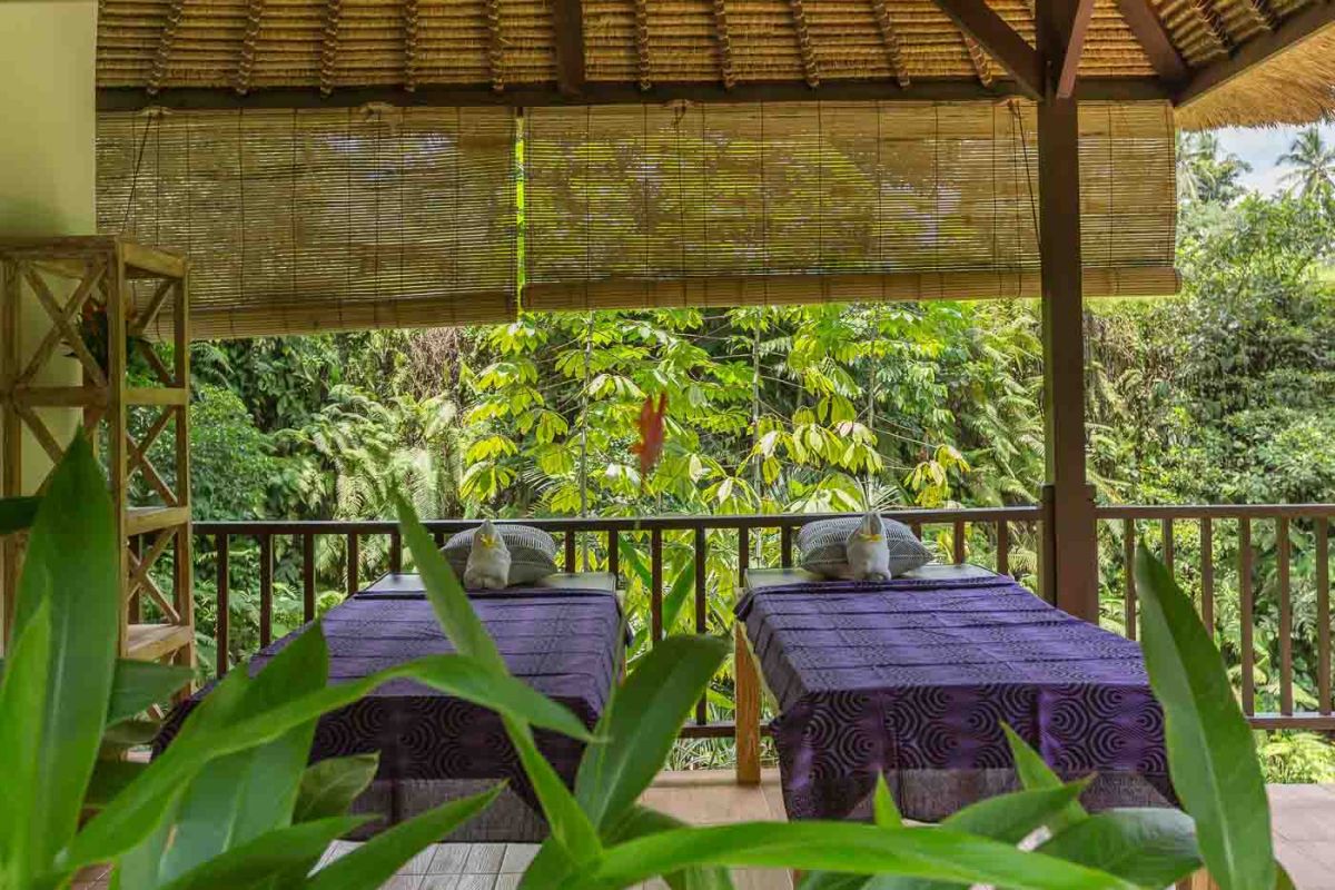 Spacious Balinese villa living room with wooden balcony, tropical furniture, stone statues, and sliding doors to lush garden. - Bali Villas