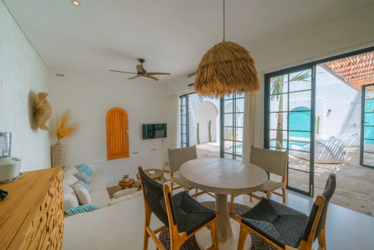 Modern tropical dining room with wooden table and chairs under a rattan pendant light, white walls, and large windows. - Bali Villas