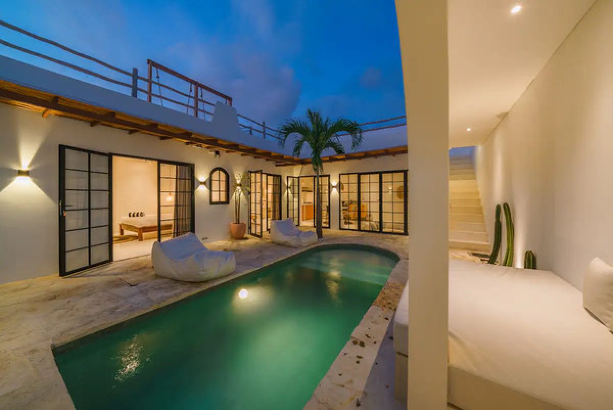 Modern tropical dining room with wooden table and chairs under a rattan pendant light, white walls, and large windows. - Bali Villas