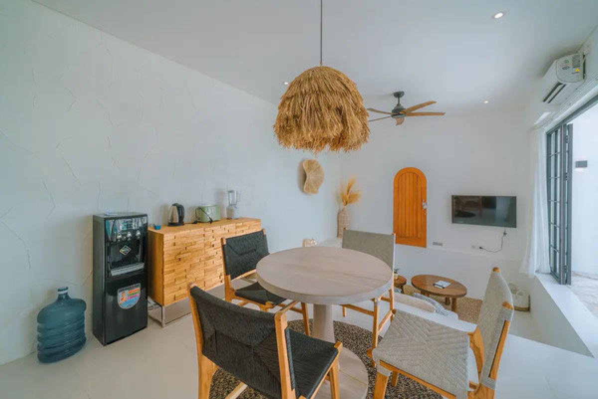 Modern tropical dining room with wooden table and chairs under a rattan pendant light, white walls, and large windows. - Bali Villas