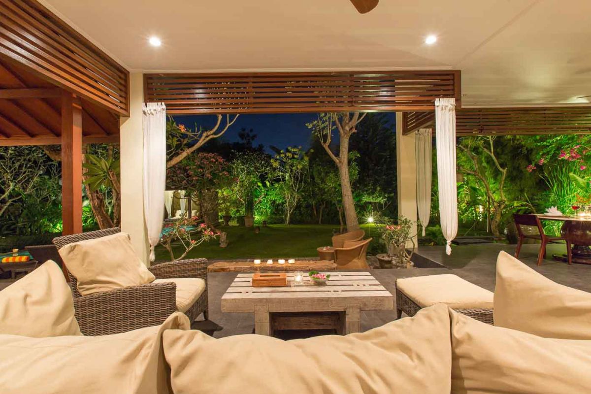 Luxurious Balinese-style bedroom suite with ornate wooden bed, beige sofa, chandelier, and tropical decor in a light, open space. - Bali Villas