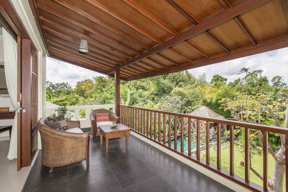 Luxurious Balinese-style bedroom suite with ornate wooden bed, beige sofa, chandelier, and tropical decor in a light, open space. - Bali Villas