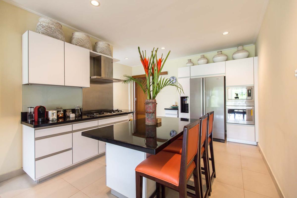 Luxurious Balinese-style bedroom suite with ornate wooden bed, beige sofa, chandelier, and tropical decor in a light, open space. - Bali Villas