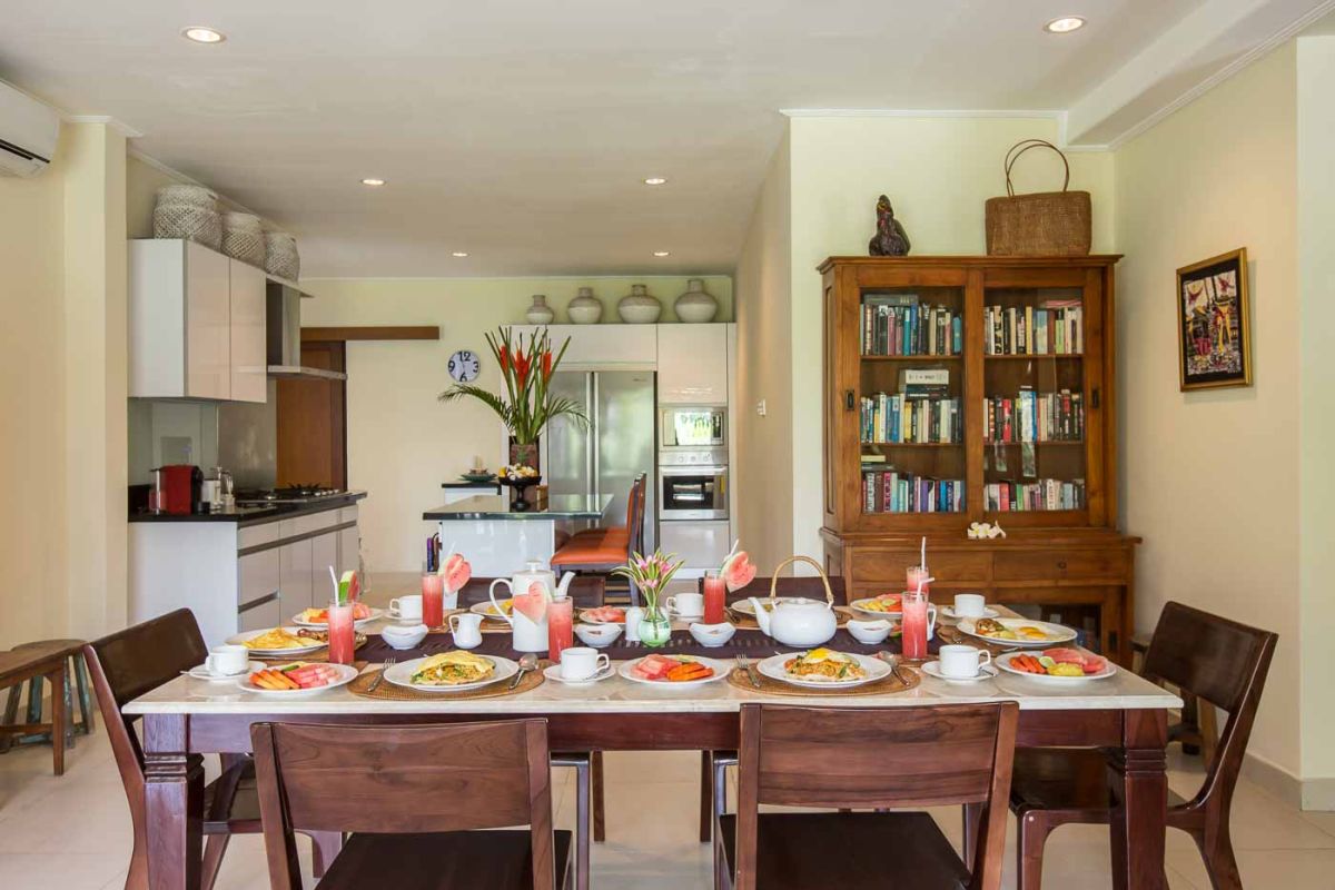 Luxurious Balinese-style bedroom suite with ornate wooden bed, beige sofa, chandelier, and tropical decor in a light, open space. - Bali Villas