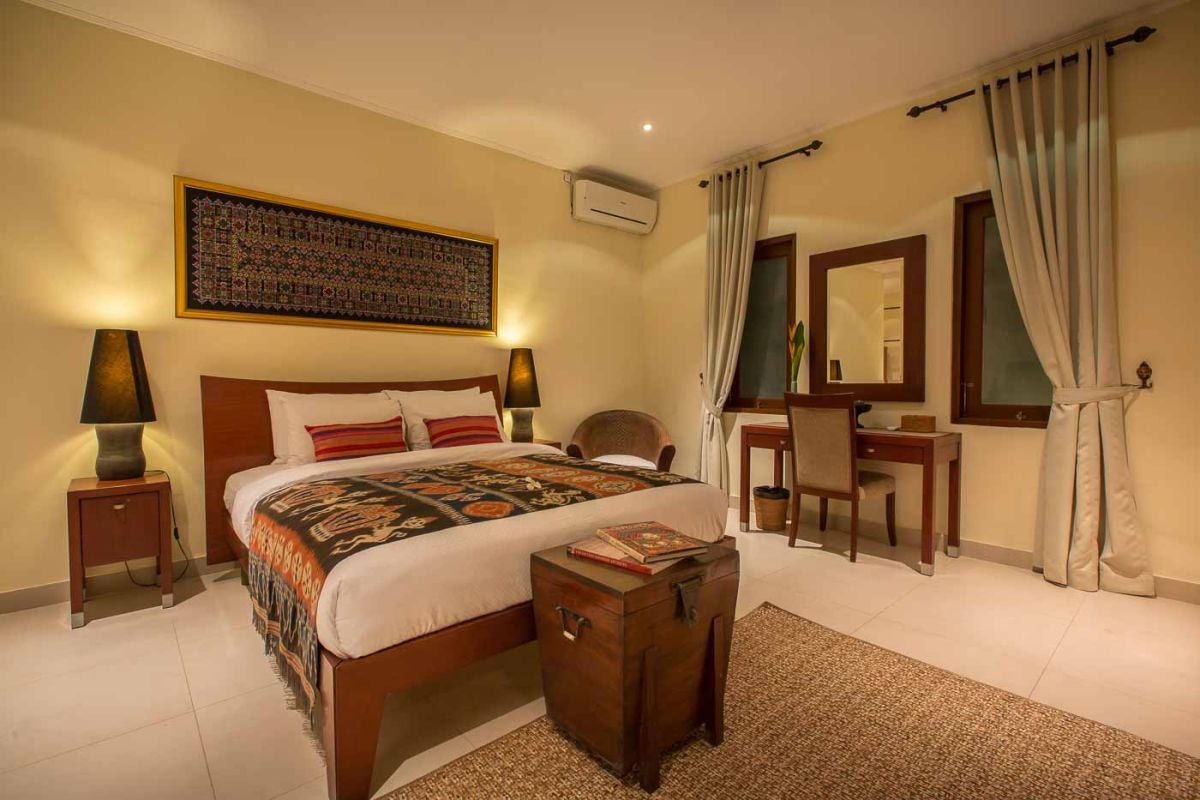 Luxurious Balinese-style bedroom suite with ornate wooden bed, beige sofa, chandelier, and tropical decor in a light, open space. - Bali Villas