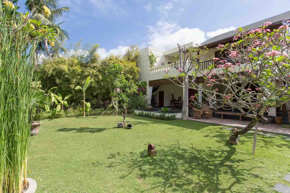 Luxurious Balinese-style bedroom suite with ornate wooden bed, beige sofa, chandelier, and tropical decor in a light, open space. - Bali Villas