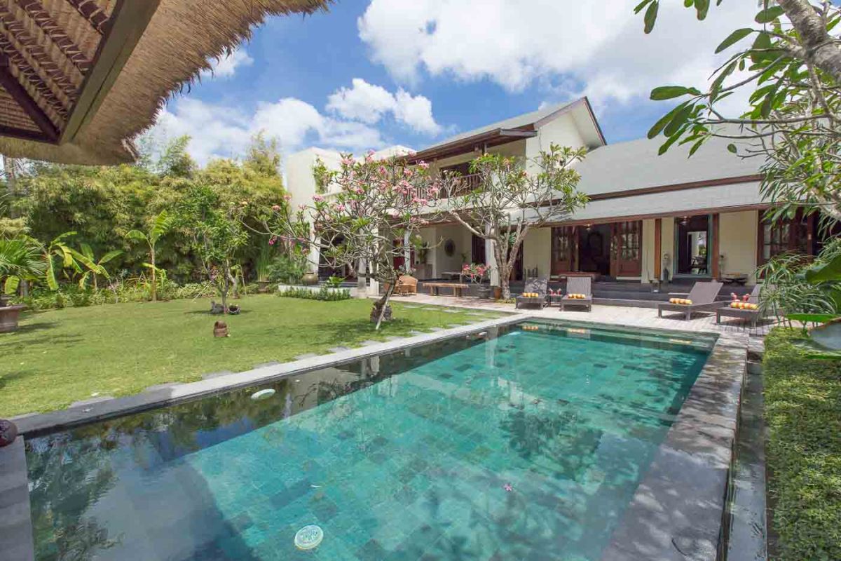 Modern Balinese villa with infinity pool, lush tropical garden, frangipani trees, and poolside loungers under sunny skies. - Bali Villas