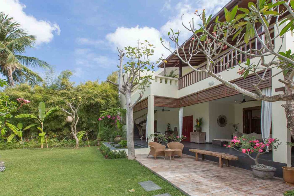Luxurious Balinese-style bedroom suite with ornate wooden bed, beige sofa, chandelier, and tropical decor in a light, open space. - Bali Villas