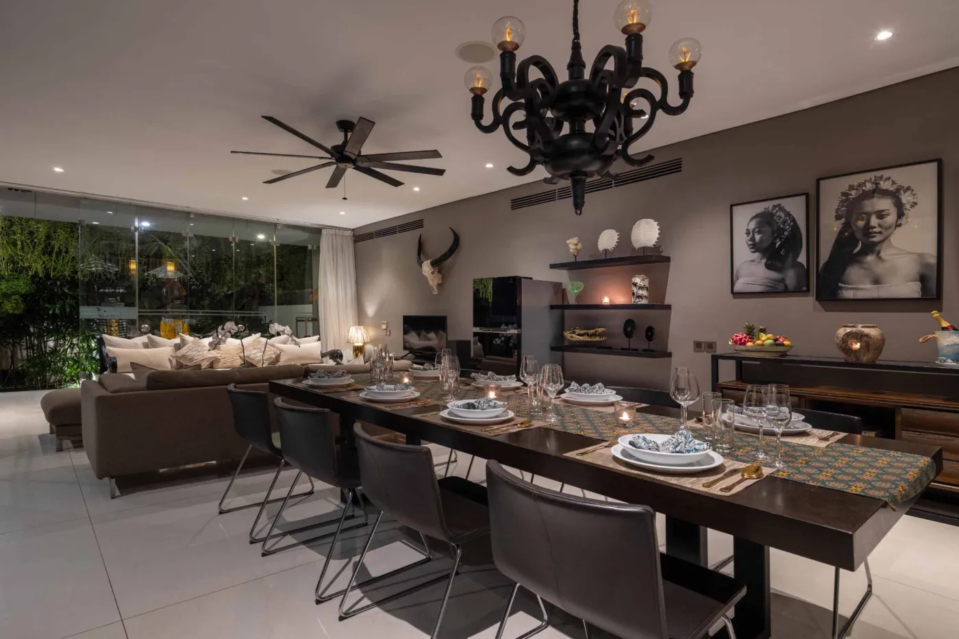 Modern dining room in a tropical villa with a set wooden table, chandelier, and glass walls overlooking a lit infinity pool and garden at night. - Bali Villas